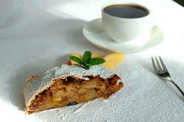 Homemade German apple strudel decorated with powered sugar, thin slice fresh apples and mint leaves. A cup of black coffee is behind and a dessert fork is in the right side.