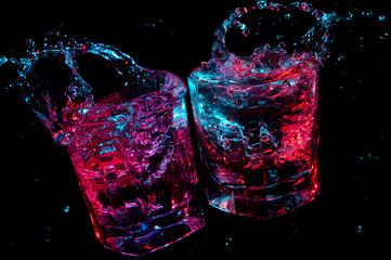 Toast of two rocks glasses with splashing liquid under colored lights isolated on a black background