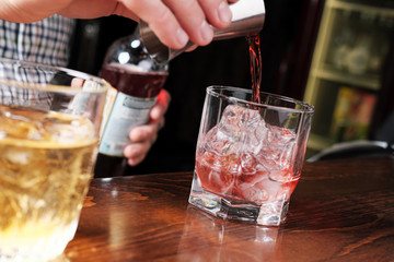 Close up shot of bartender hand pouring drink from measuring cup into a cocktail glass filled with ice cubes. Barman preparing fresh negroni cocktail