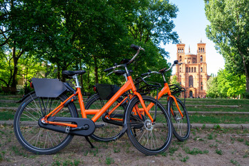 Fototapeta na wymiar Two orange bicycles parked towards St. Thomas protestant church (Thomaskirche) in Berlin, Germany. Urban bike share concept for weekend gateaways and sightseeing eco friendly city cycle tours.