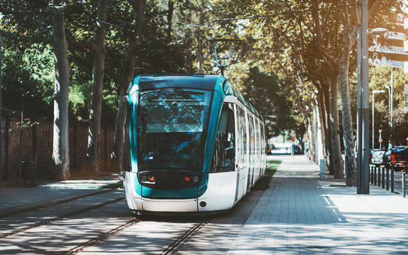 A modern neat tram in city alleyway surrounded by trees; blue and white city streetcar near the sidewalk made of pavement stones; contemporary tramcar on the station with the road on the right
