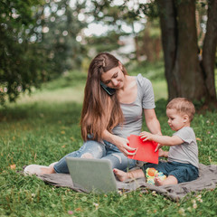 Mother with son sitting in the park, using phone and work on laptop.