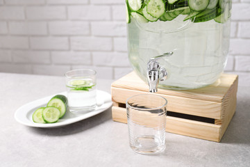 Jar dispenser of fresh cucumber water and glasses on table against brick wall. Space for text