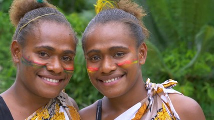 SLOW MOTION, PORTRAIT, CLOSE UP: Beautiful girls wearing traditional face paint and native outfits before a Melanesian ceremony. Lovely black friends wear vibrant make up and smile at the camera.