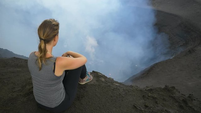 CLOSE UP: Young traveler woman sits on the crater rim of stunning Mount Yasur in Vanuatu. Unrecognizable female tourist observing the smoke and ash rising from the active volcano roaring below her.