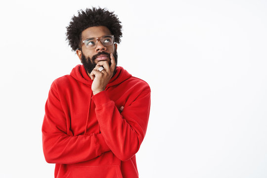 Creative african american bearded guy with afro hairstyle in glasses and red hoodie creating new song, standing in thoughtful pose touching chin looking dreamy, focused at upper right corner, thinking