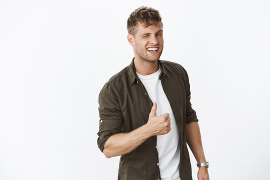 Hey you doing awesome. Portrait of friendly enthusiastic handsome fair-haired bearded guy winking at you and showing thumbs up in like and approval, cheering being supportive over grey wall