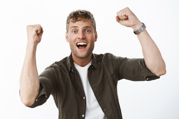 Fototapeta premium Optimistic and excited charismatic blond guy with blue eyes and white broad smile raising clenched fists in triumph, support and cheer smiling delighted from success and victory over gray wall