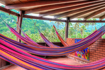 A relaxing peaceful spot with multiple colorful hammocks in a wooden hut in Tyrona, Colombia. The background shows green mountains with palm trees and tropical plants. Tyrona National Park, Colombia.