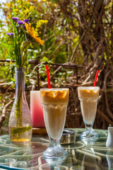 Two Ice coffee glasses with red drinking straw, vase with flowers, ashtray and red candle on the metal glass table surrounded by exotic plants. Romantic place.