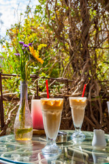 Two Ice coffee glasses with red drinking straw, vase with flowers, ashtray and red candle on the metal glass table surrounded by exotic plants. Romantic place.