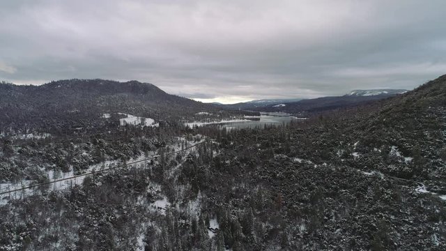 Aerial shot flying over snowy landscape towards a lake. Road on the side, during overcast day. Mountains in distance.