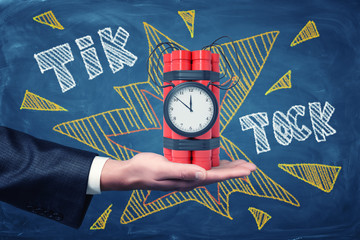 Man's hand facing up holding dynamite bundle with time bomb on blackboard background with chalk...
