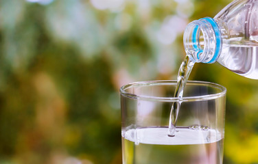 Pouring of clear drink water from bottle into the glass on blurred green nature background with copy space for text.