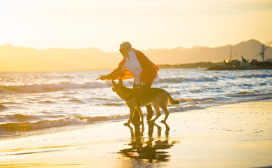 Happy attractive senior woman with her german shepard dog walking on the beach at autumn sunset