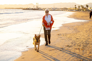 Happy attractive senior woman with her german shepard dog walking on the beach at autumn sunset