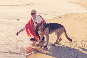 Happy attractive senior woman with her german shepard dog playing on the beach at autumn sunset