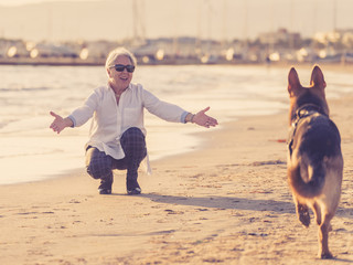 Happy attractive senior woman with her german shepard dog playing on the beach at autumn sunset