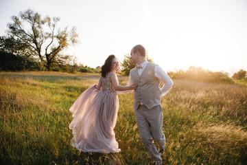 Fototapeta na wymiar Wedding near the river in field at sunset with brown horse. Bride in light airy dress in color of dusty rose. Beige dress with sparkles. Light suit with bow tie. bride and groom embrace and kiss.