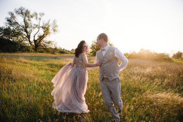 Wedding near the river in field at sunset with brown horse. Bride in light airy dress in color of dusty rose. Beige dress with sparkles. Light suit with bow tie. bride and groom embrace and kiss.