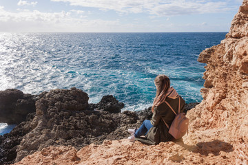 Happy female tourist in coat sits on cliff and enjoys sea view in Malta