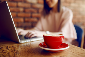 Selective focus on cup of delicious coffee. In background woman using laptop.