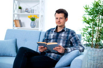 Handsome young man guy reading a book sitting on the couch in his living room