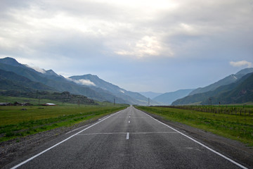 Chui tract is the main road of Altai. Straight long road extending into the distance towards the mountains. Along the edges of the road are green fields and farms. The sky is gray and gloomy