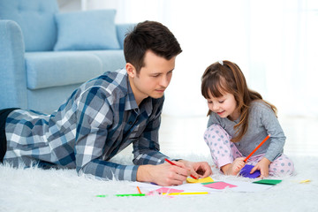 father and little daughter having quality family time together at home. dad with girl lying on warm floor drawing with colorful felt tip pencils.