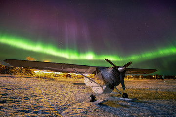 Aurora borealis over a small airplane parked on a frozen lake during an arctic night.