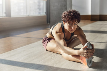 Young motivated woman wearing activewear sports-bra, sneakers in gym stretching foot excercising, working-out, sitting on floor enjoying pleasant pain in muscles after productive training session