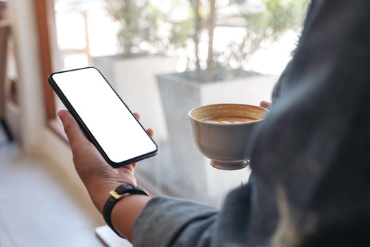 Mockup image of hands holding black mobile phone with blank desktop screen while drinking coffee in cafe
