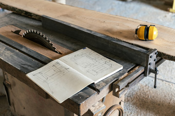 Design wood small business concept. Technical drawing in notebook lying on top of wood in garage or workroom near yellow headset and table saw