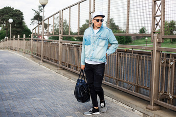 Stylish cute guy is standing on the bridge, wearing a denim jacket cap, listening to music through headphones free hands