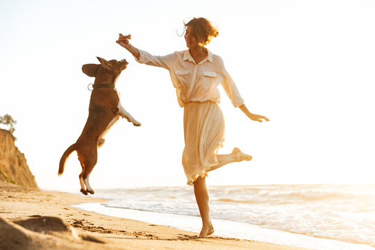 Image of happy woman 20s playing with her dog, while walking by seaside
