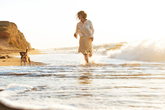 Image of lovely woman 20s running with her dog, by seaside in the morning