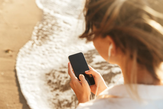 Image of caucasian girl 20s wearing earphones using mobile phone, while walking by seaside