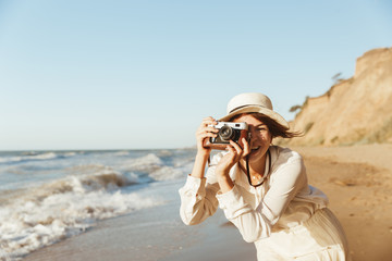 Image of cheerful woman 20s photographing on retro camera, while walking by seaside