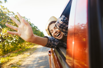 Image of caucasian woman 20s wearing straw hat laughing and waving hand out of the window, while riding in car