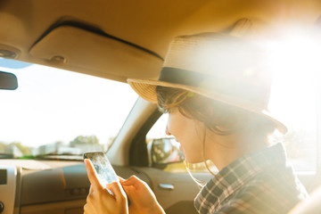 Fototapeta na wymiar Image of young woman 20s wearing straw hat using mobile phone, while riding in car