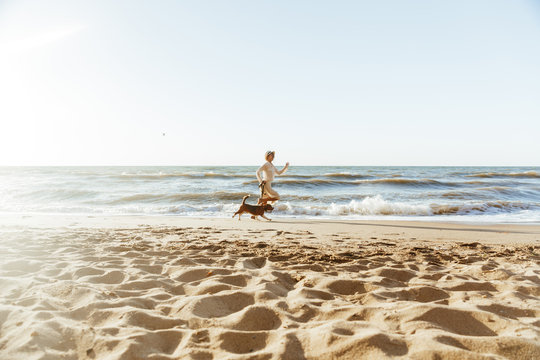 Image of positive woman in straw hat, running with her brown dog along the coast