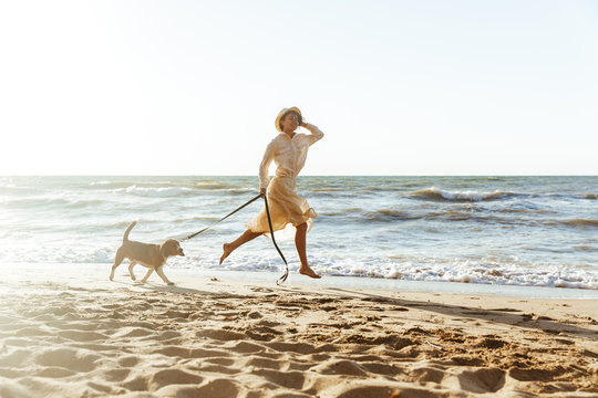Image of caucasian woman in straw hat, running with her brown dog along the coast