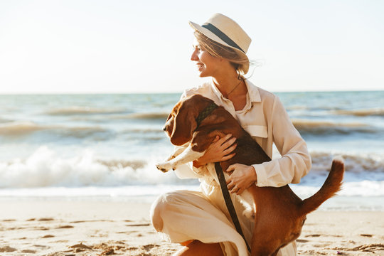 Image of pretty woman in straw hat hugging her brown dog, while walking by seaside