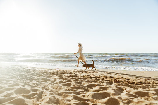 Image of happy woman in straw hat, running with her brown dog along the coast