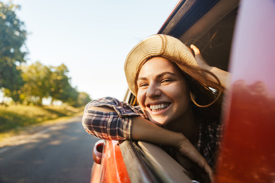 Image of joyous woman 20s wearing straw hat smiling and looking out of the window, while riding in car