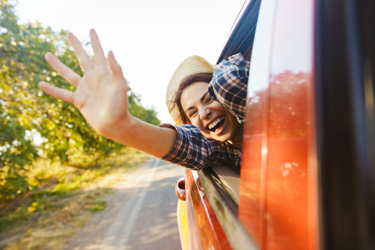 Image of gorgeous woman 20s wearing straw hat laughing and waving hand out of the window, while riding in car