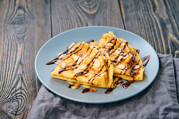 Two pancakes with chocolate syrup, almond flakes on plate, honey flows from spoon, side view