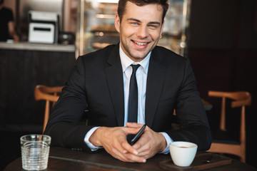 Handsome young businessman in formal clothes indoors in cafe using mobile phone.