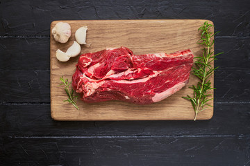 Fresh raw beef meat with garlic and rosemary on cutting board on black wooden table. Copy space, top view.