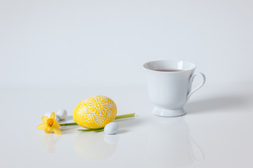 Cup of tea with yellow painted egg and daffodil on white background. Copy space, selective focus. Spring concept. Holiday morning concept.
